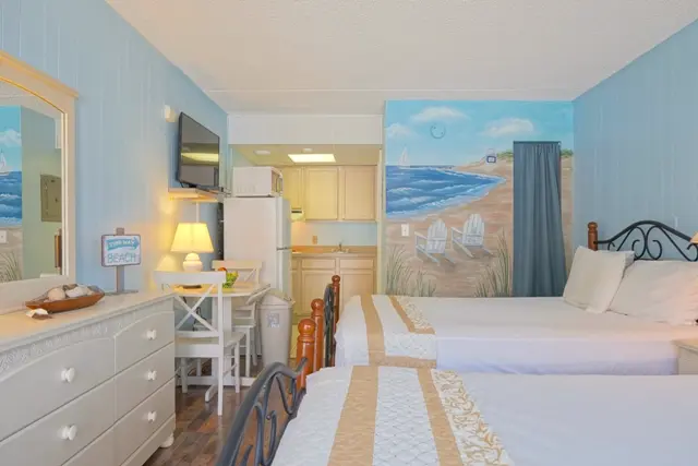 A studio with two queen beds, a white dresser, a beach mural on the wall, and the kitchenette in the back
